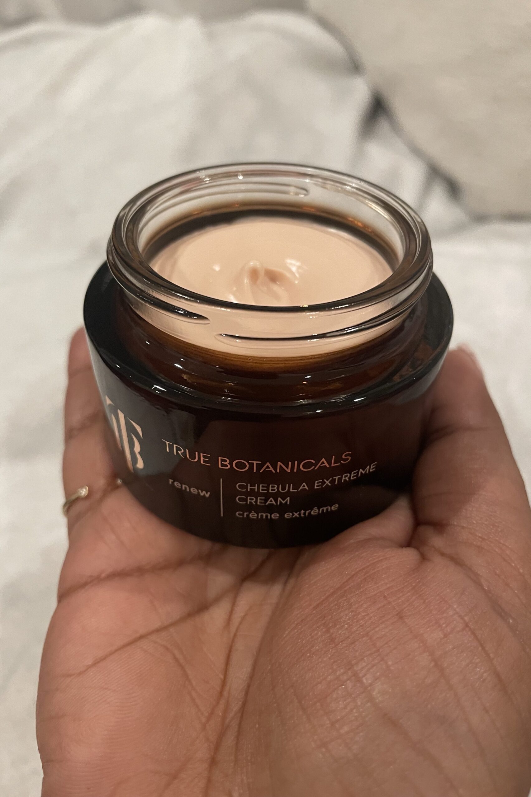 A person holding a jar of true botanicals cream on a bed.
