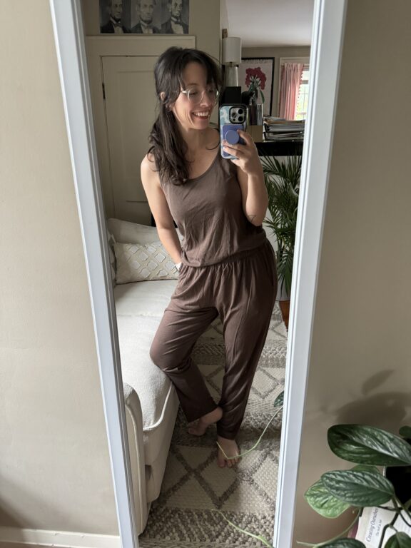 Woman wearing brown loungewear stands in front of a full-length mirror taking a selfie in a living room with plants and framed photos on the wall.