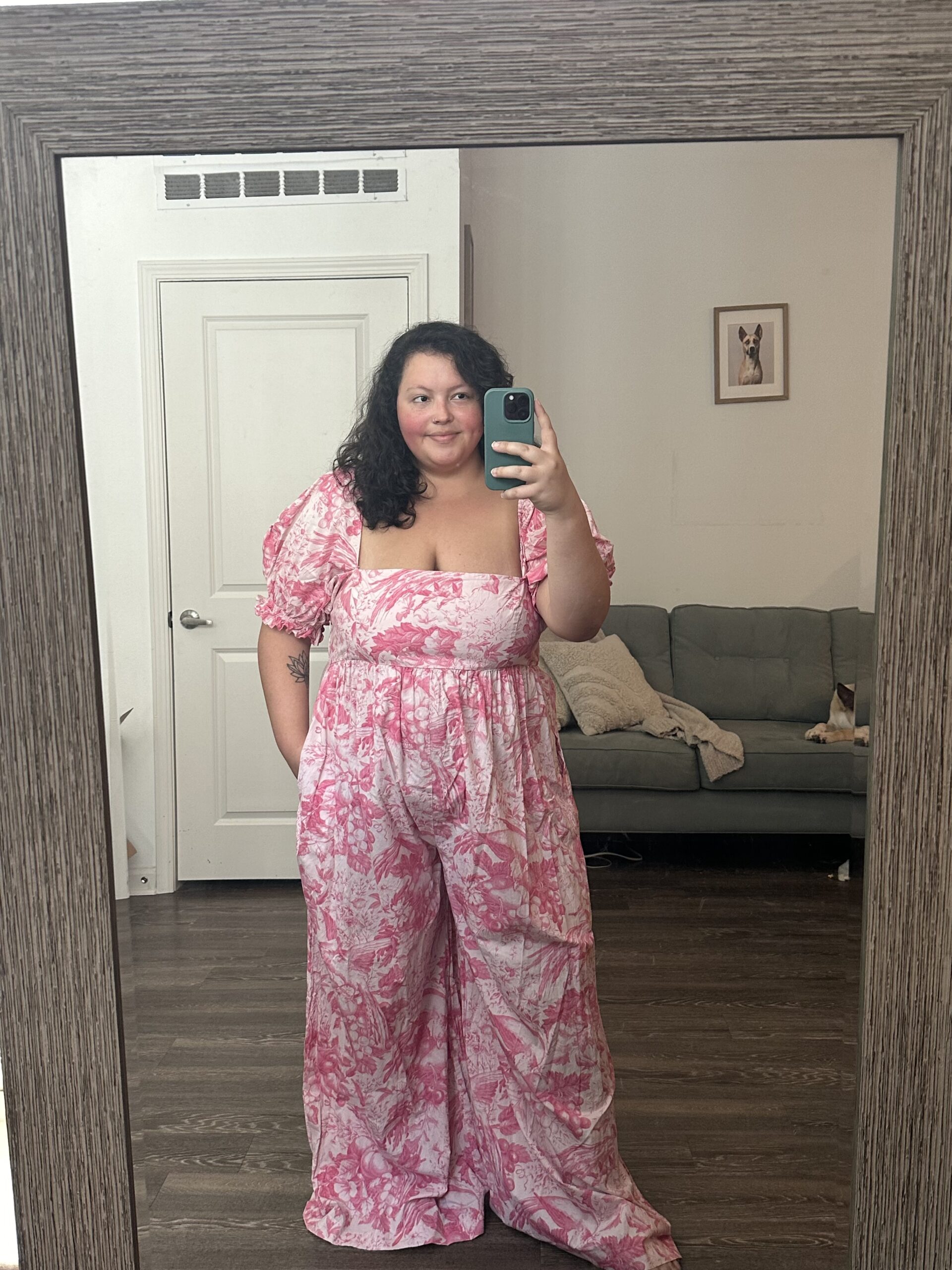 Person takes a mirror selfie in a room, wearing a pink and white floral jumpsuit. A couch with a blanket and a framed picture are visible in the background.