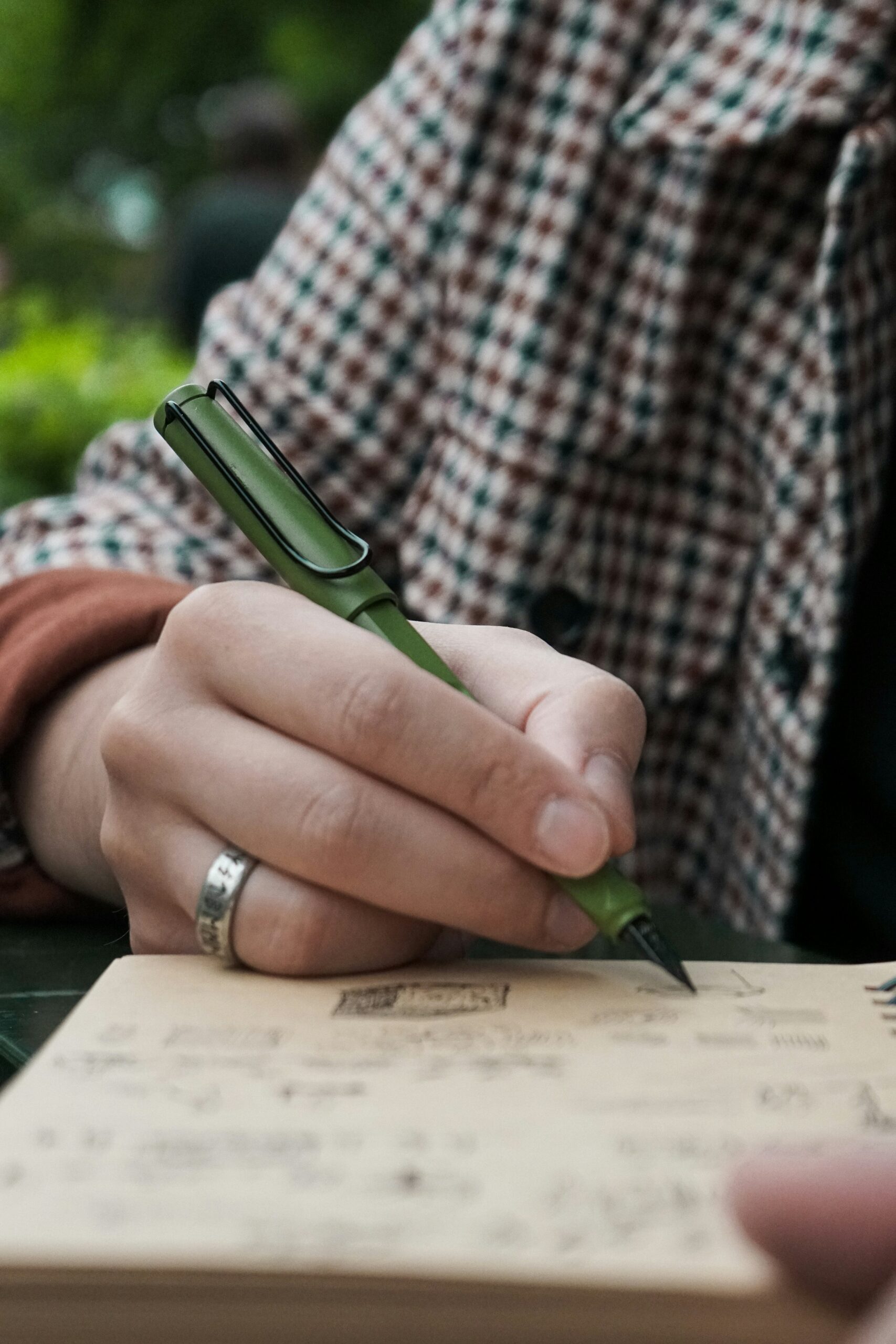 A person wearing a checkered jacket and a silver ring writes in a notebook with a green fountain pen.