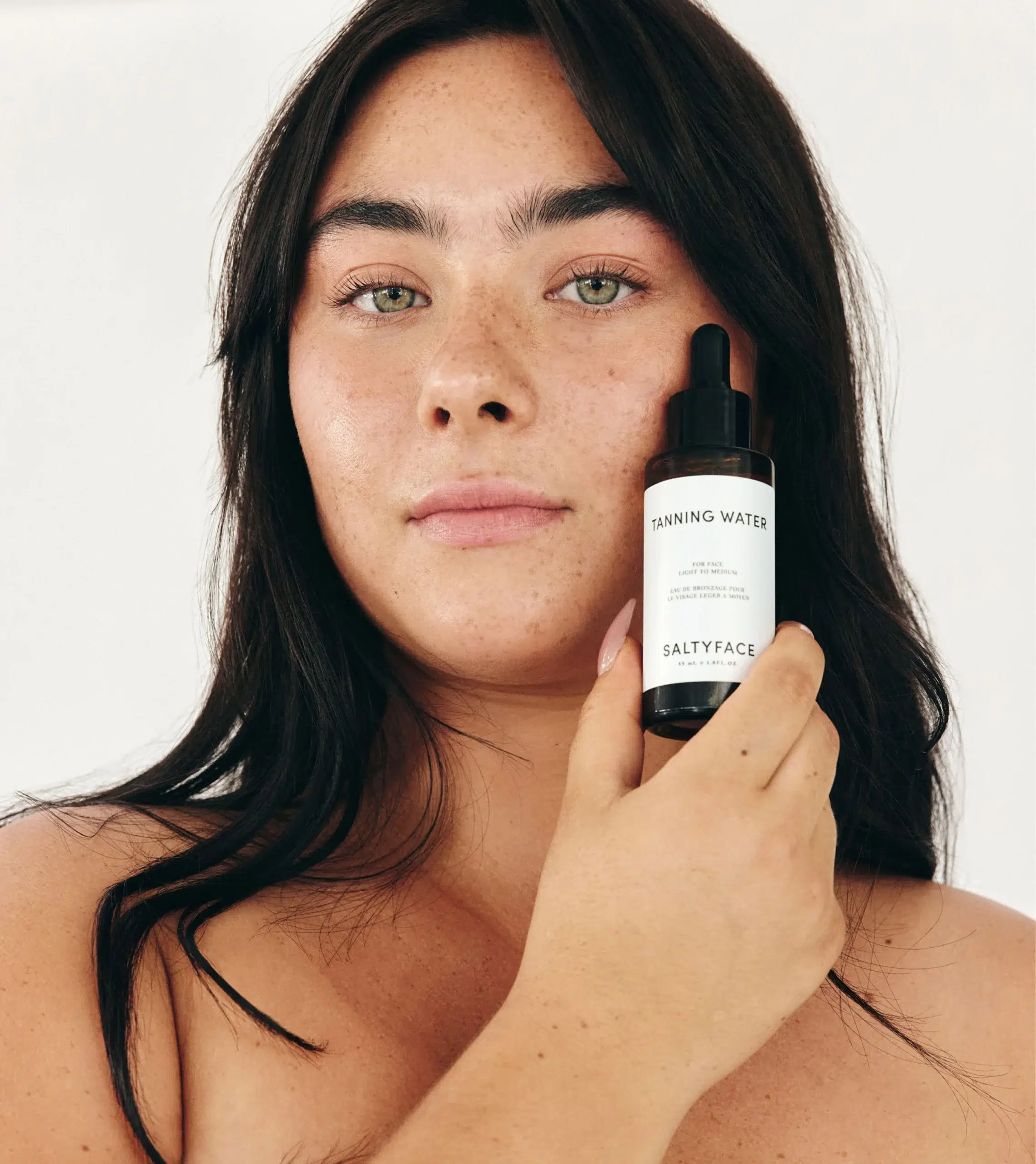 Person holding a bottle of Saltyface Tanning Water close to their face, with a neutral expression against a plain background.