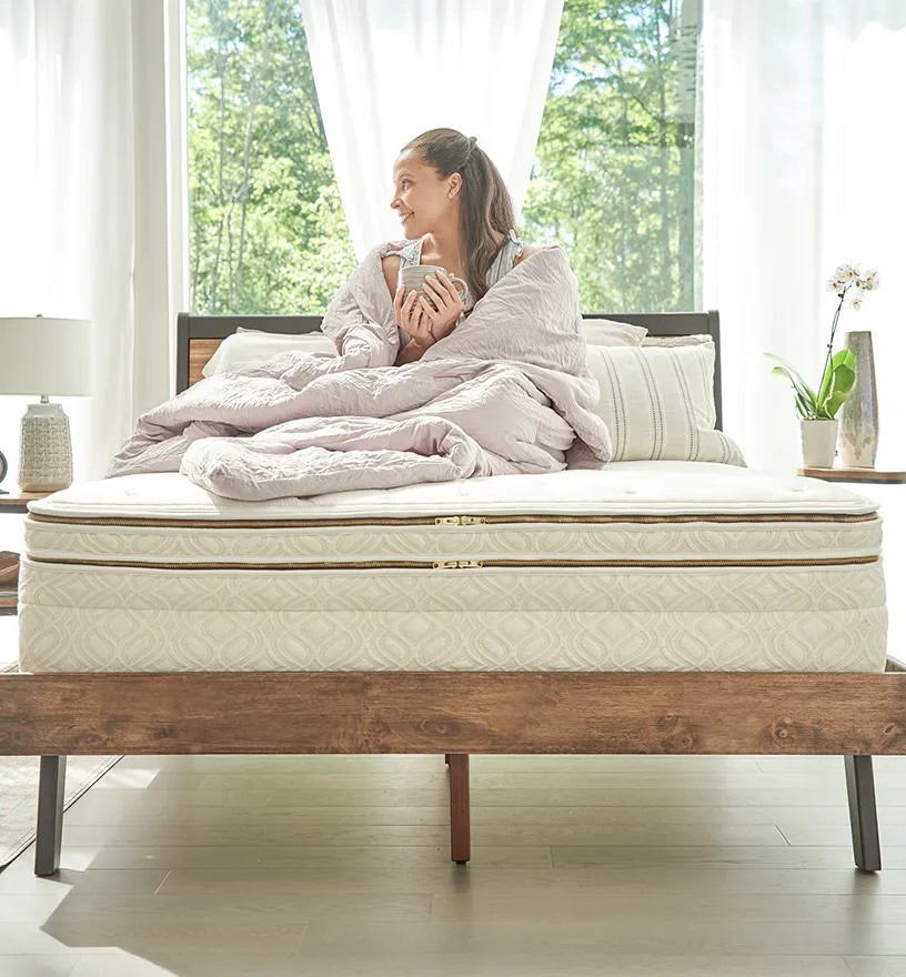 A person sits on a Naturepedic mattress with a light comforter, holding a mug, in a sunlit room. Large windows offer a view of trees outside.