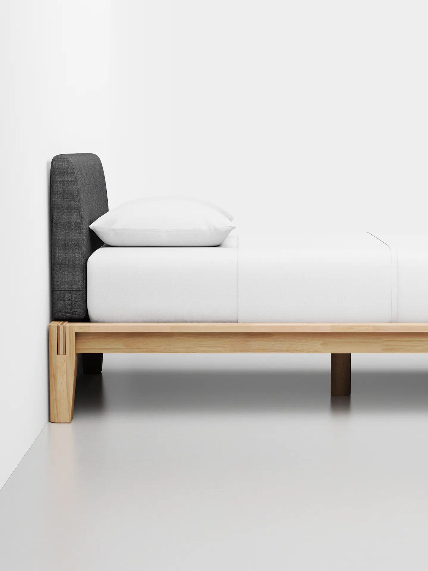 A minimalist bed with a wooden frame and white bedding, positioned against a white wall. The bed has a dark grey upholstered headboard.