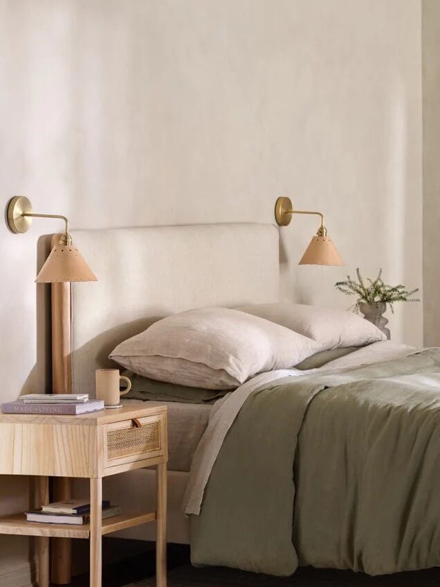 A bedroom with a neatly made bed featuring green and beige bedding, a wooden nightstand with a book and cup, wall-mounted lamps, and a mirror in the background. Text reads: "FEATURED: COTTON LINEN OAT, MAPLE WOOD.