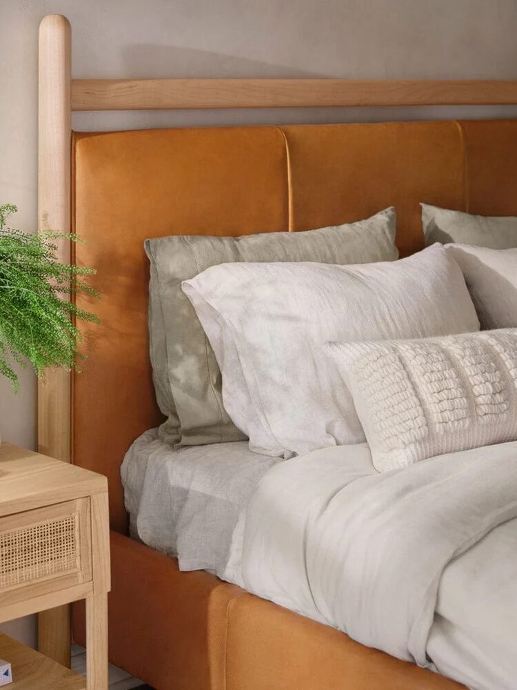 A neatly made bed with white and beige linens and pillows is next to a wooden nightstand holding a green plant, a lamp, and stacked books.