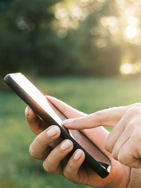 A person using a smartphone outdoors in a grassy area with sunlight shining through the trees.