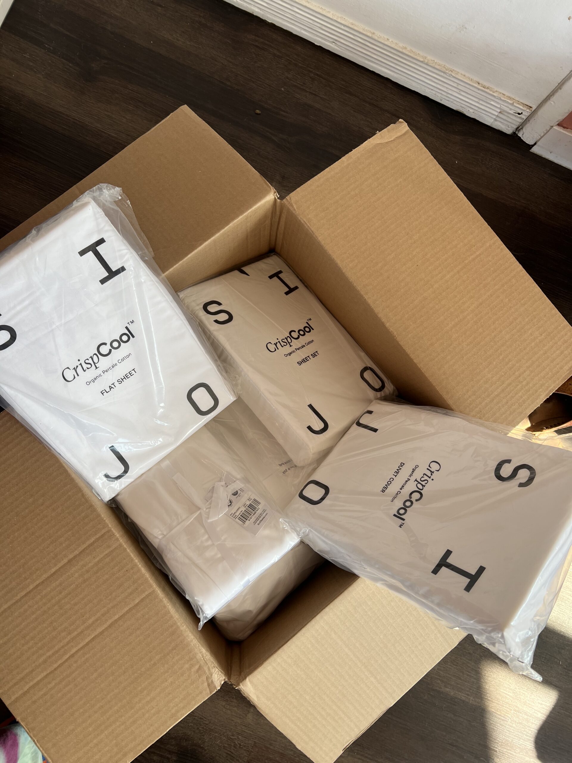 An open cardboard box containing several packaged white bed sheets labeled "CrispCool.