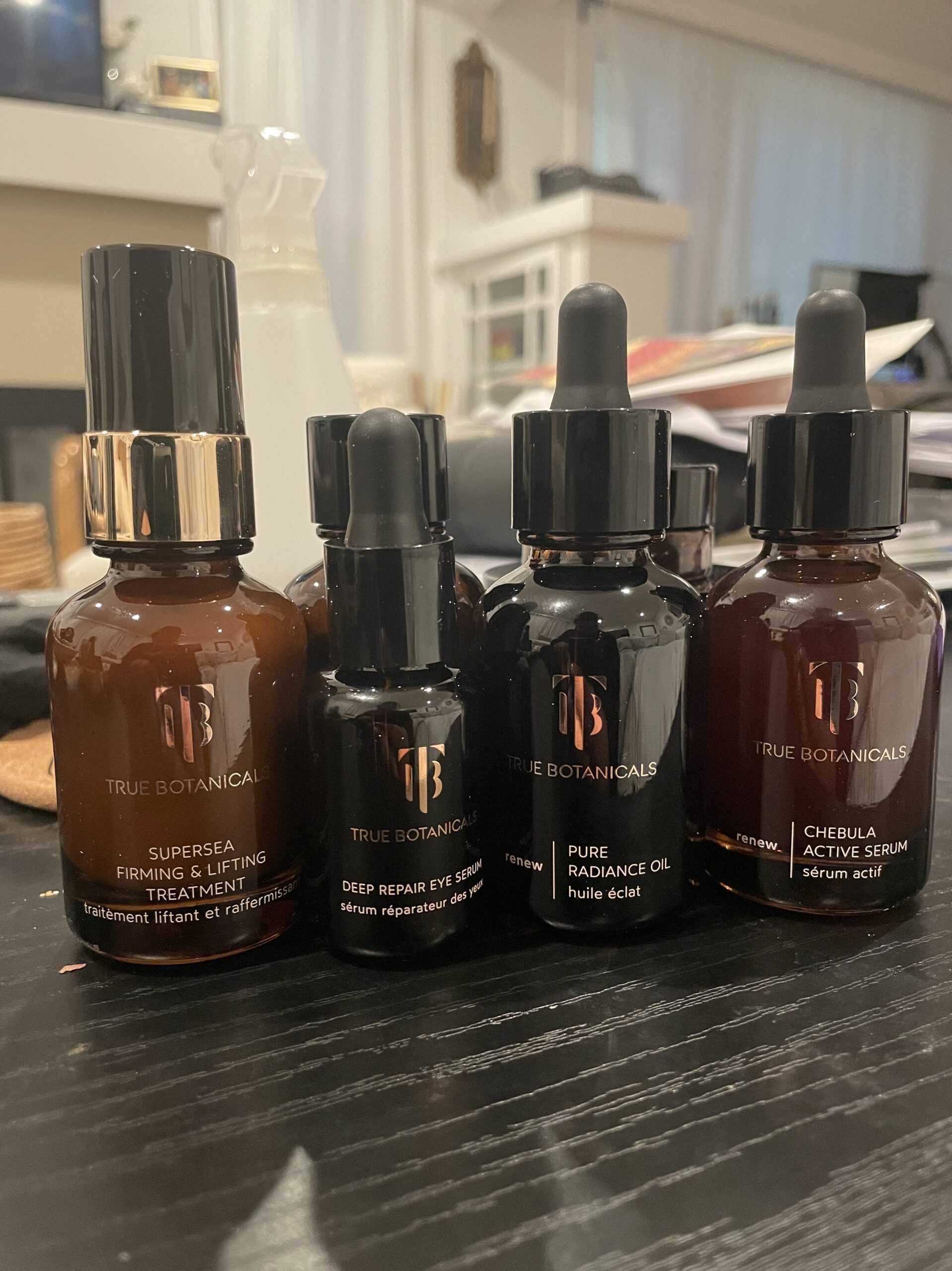 A collection of five True Botanicals skincare products, including serums and treatment bottles, arranged on a dark surface with a blurred background.
