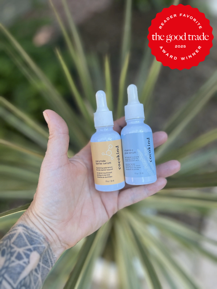 A tattooed hand holding two bottles of eco-oil over blurred greenery. One bottle is labeled "Calm Down" and the other "Glow Brighter." A red badge reads "Reader Favorite, AlsWell Blog, 2025 Award Winner.