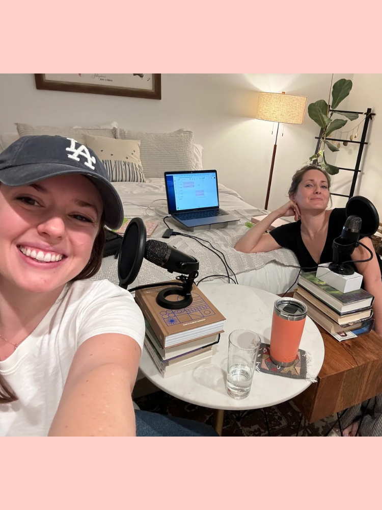 Two women sitting on a cozy couch setup, recording a podcast with microphones, a laptop, and books. One woman wears a cap and smiles; the other rests her chin on her hand.