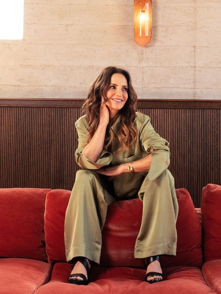 A person in a green outfit sits on a large red couch, smiling, with one hand resting on their cheek. There are two windows in the background.