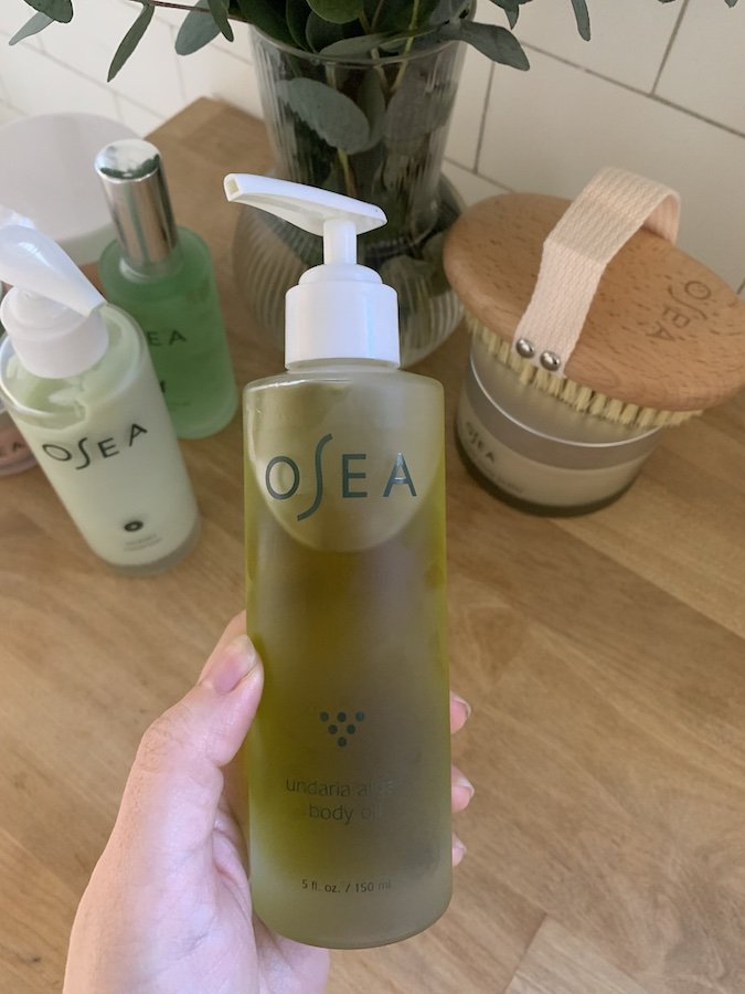 Hand holding a bottle of Osea Undaria Algae Body Oil, with other skincare products and a brush in the background on a wooden surface.