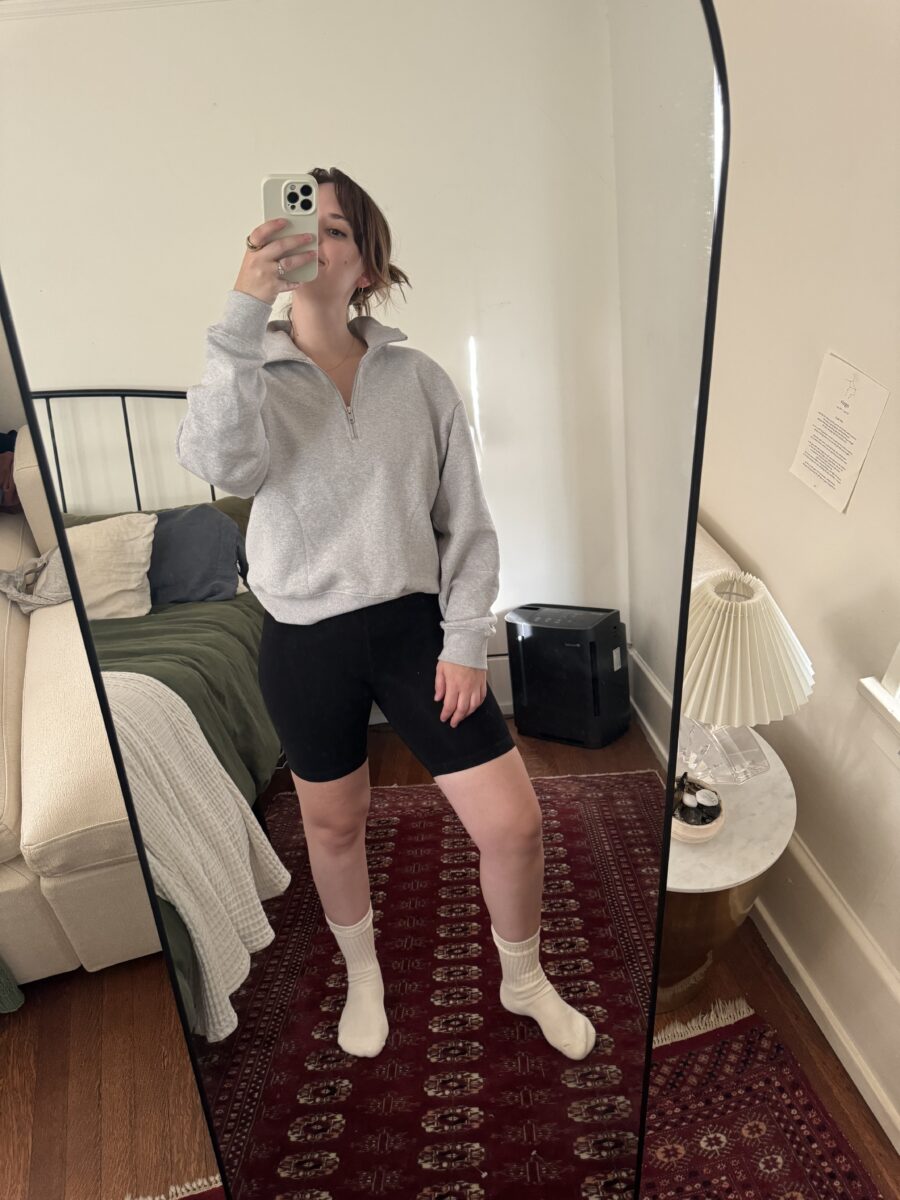 Person taking a mirror selfie in a bedroom, wearing a gray sweatshirt, black shorts, and white socks. A bed with green blankets and a side table with a lamp are visible.