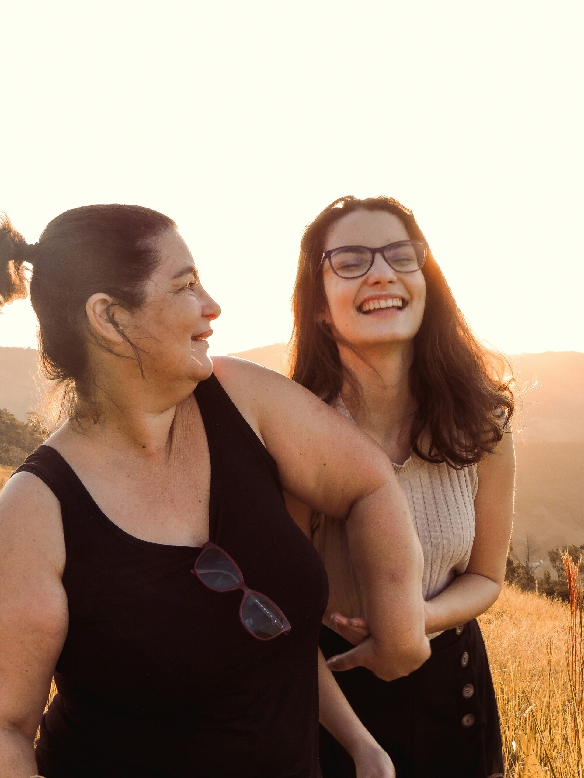 Two women stand outdoors in a field at sunset, smiling and laughing together. One wears glasses and a sleeveless top, the other has sunglasses hanging from her tank top.