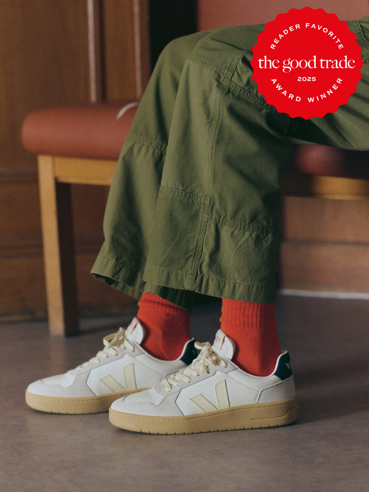 A person wearing olive green pants, red socks, and cream-colored sustainable sneakers from Veja sits on a red cushioned chair. A red "AlsWell Blog 2025 Reader Favorite Award Winner" badge is in the top right corner.