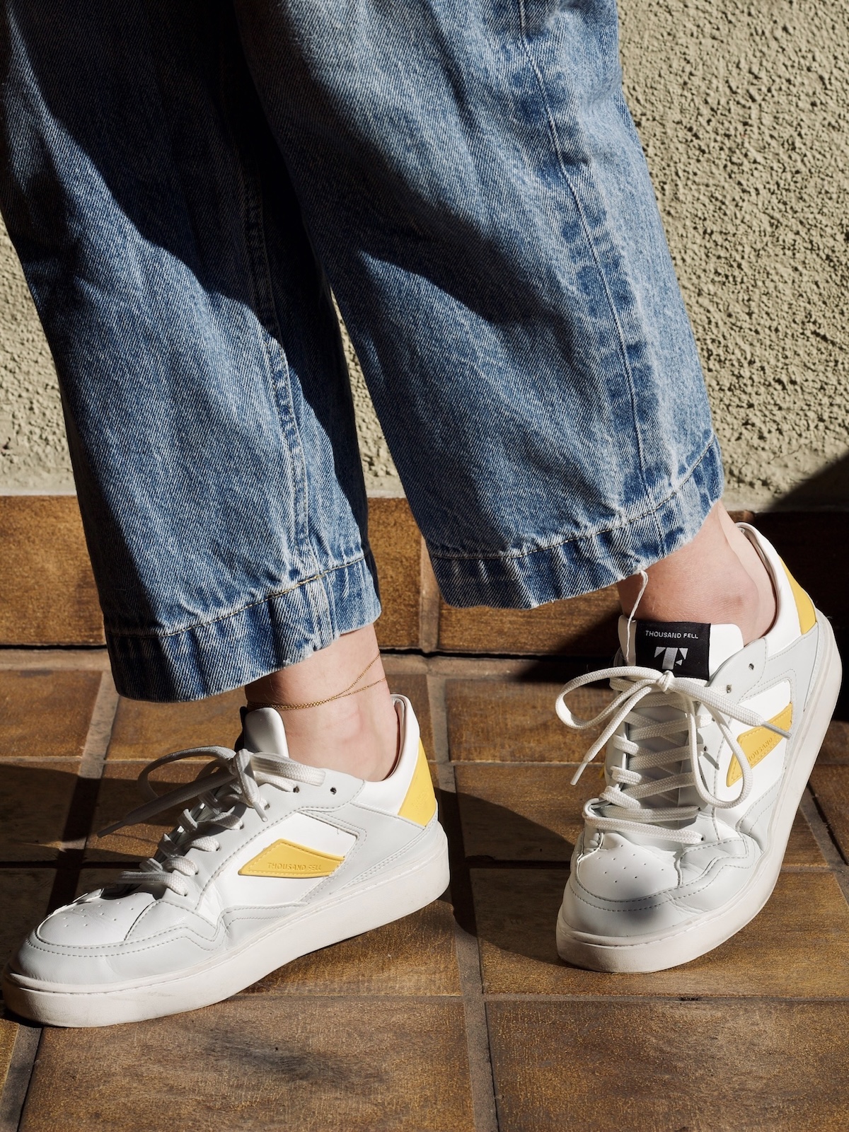 Person wearing loose blue jeans and white sustainable sneakers with yellow accents, standing on a brown tiled floor near a textured beige wall.
