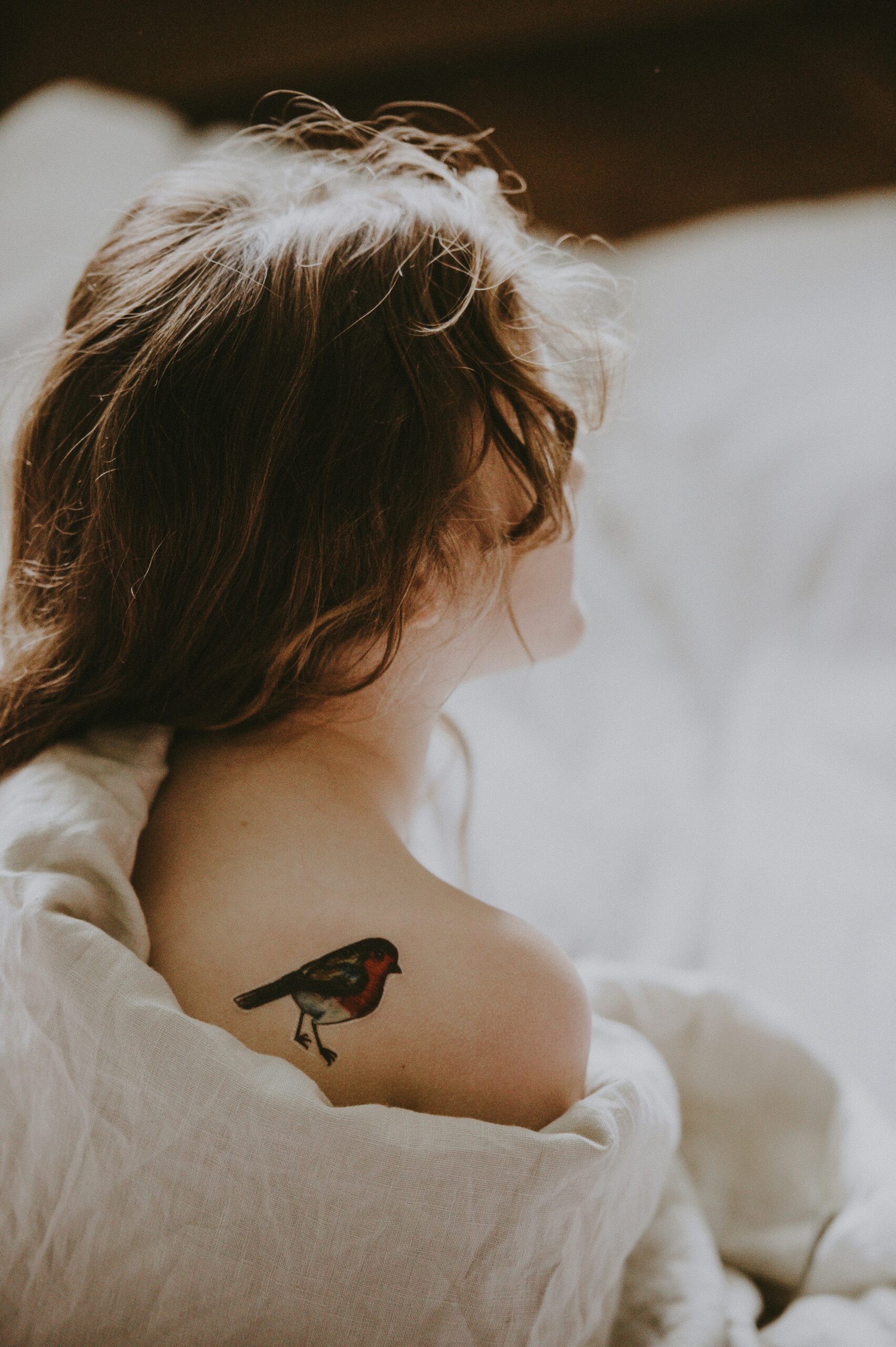 A person with long hair sits with their back turned, revealing a bird tattoo on their right shoulder. They are wrapped in a light-colored sheet.