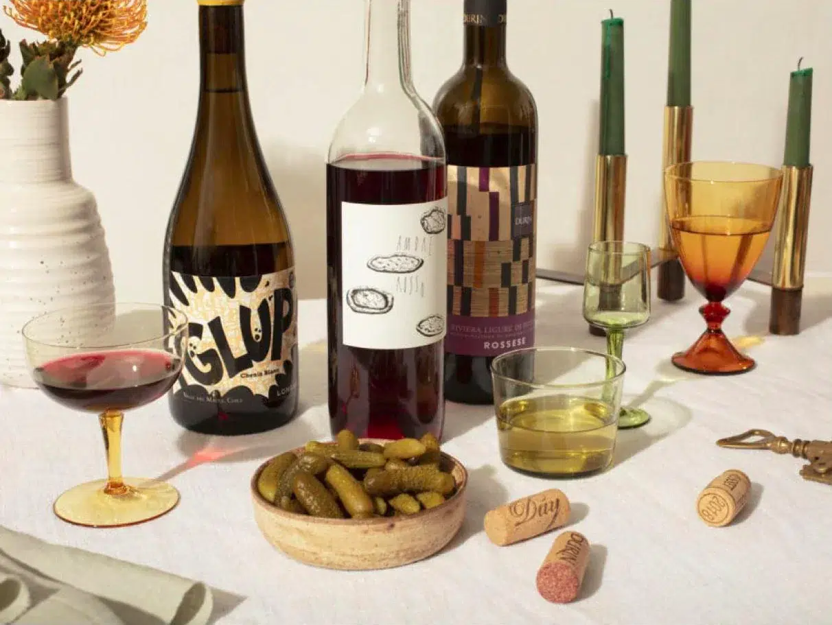 A table with three wine bottles, a bowl of pickles, various drinking glasses, and corks on a white tablecloth. Green and orange candles are in the background.