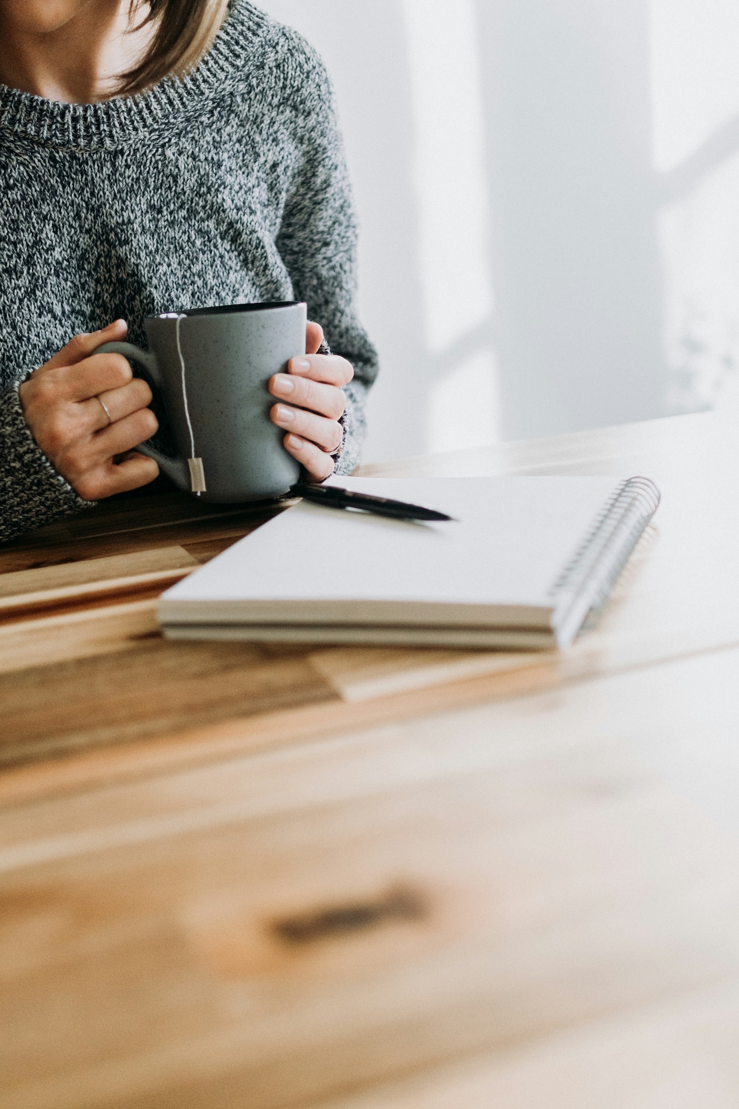 Person holding a mug, sitting at a wooden table with an open spiral notebook and pen nearby.