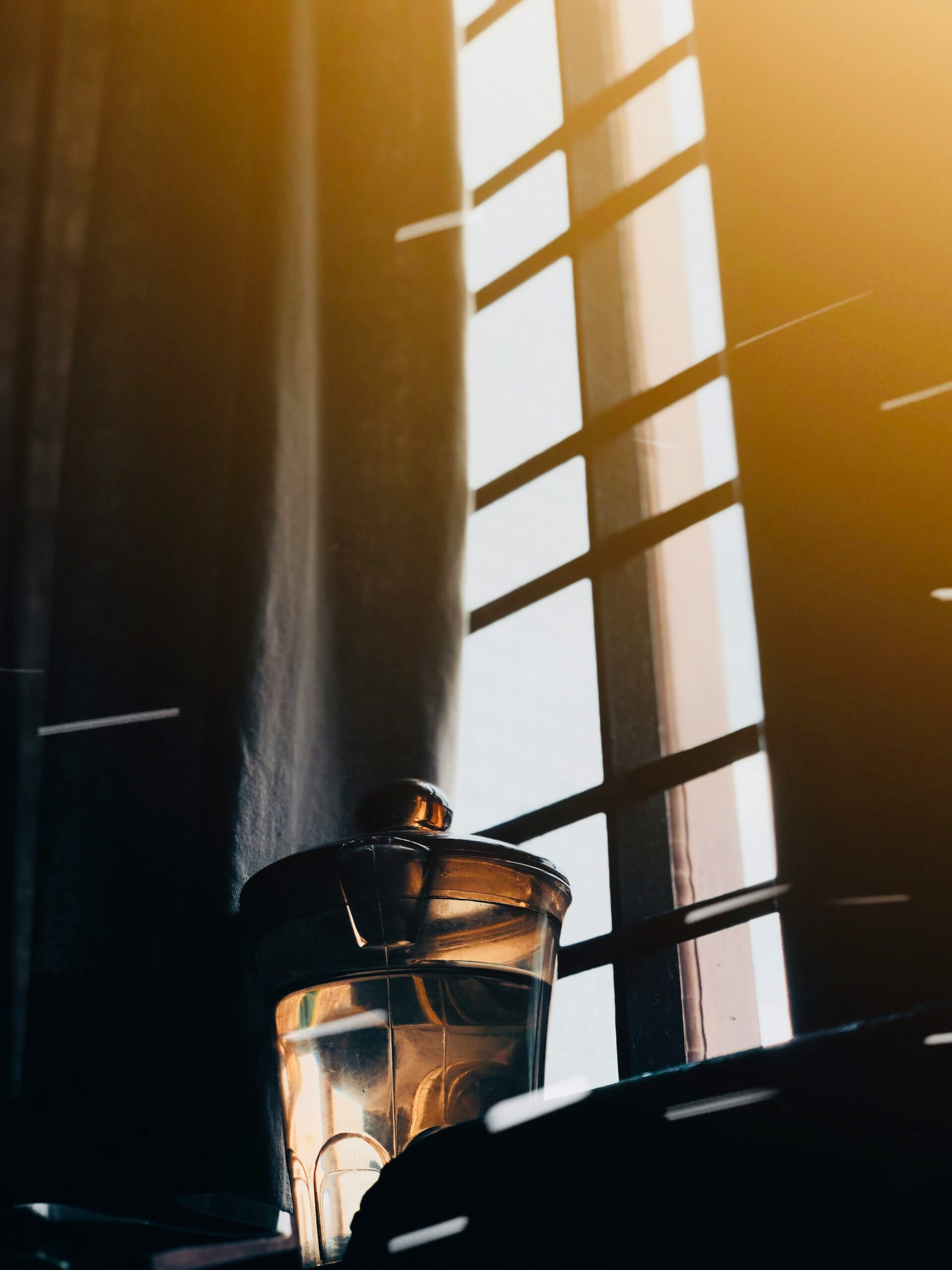 A glass jar with a lid sits by a window with a metal grid. Sunlight streams through, casting warm tones and shadows.