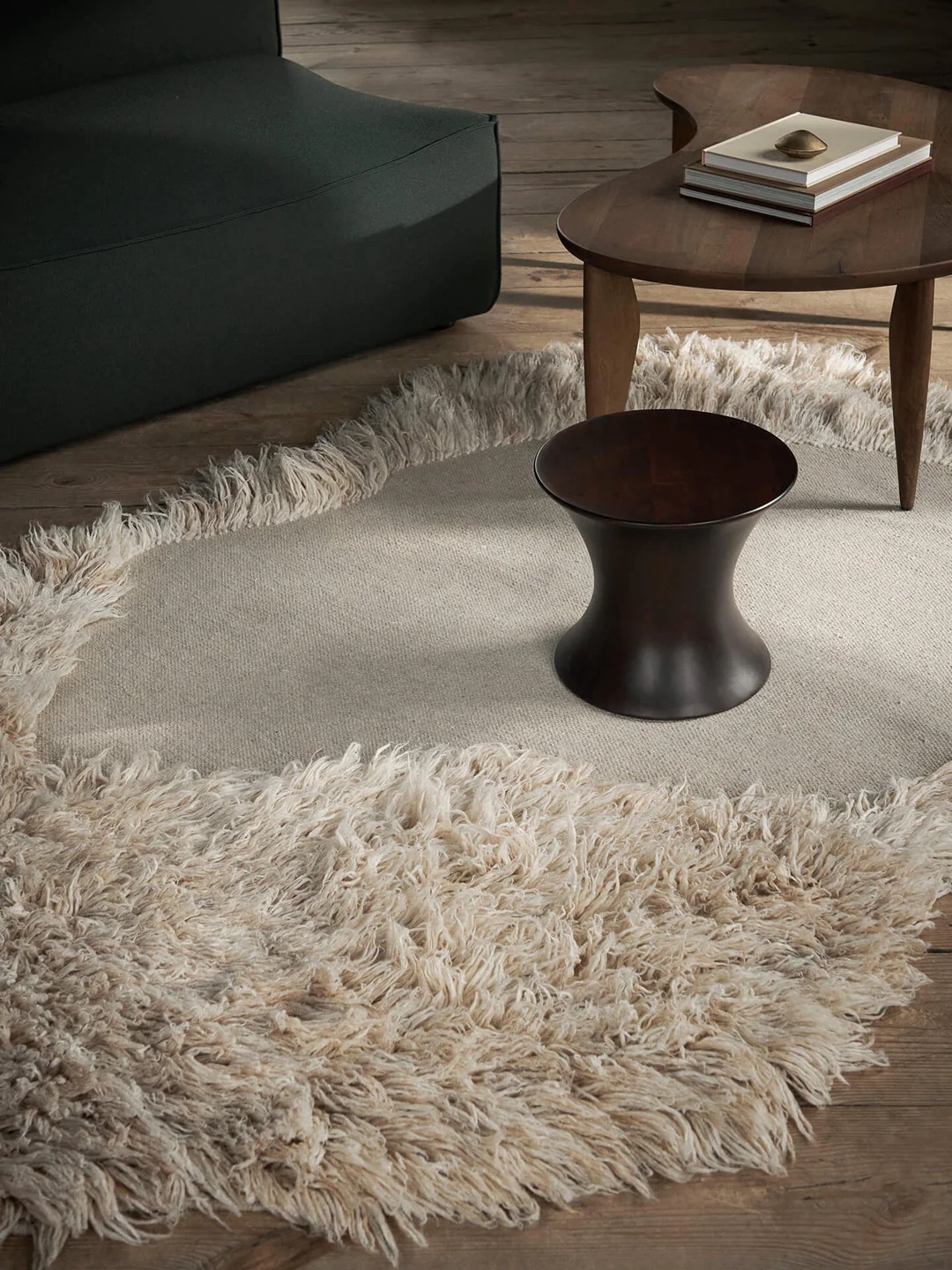 A living room scene with a shaggy beige rug, a dark wooden side table, a larger wooden coffee table with books, and a dark-colored sofa.