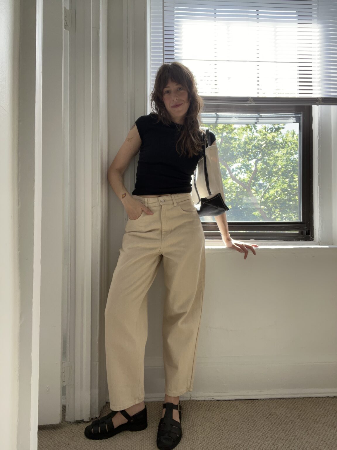 Person in a black shirt and beige pants stands casually by a window with blinds, sunlight streaming in and a green tree visible outside.