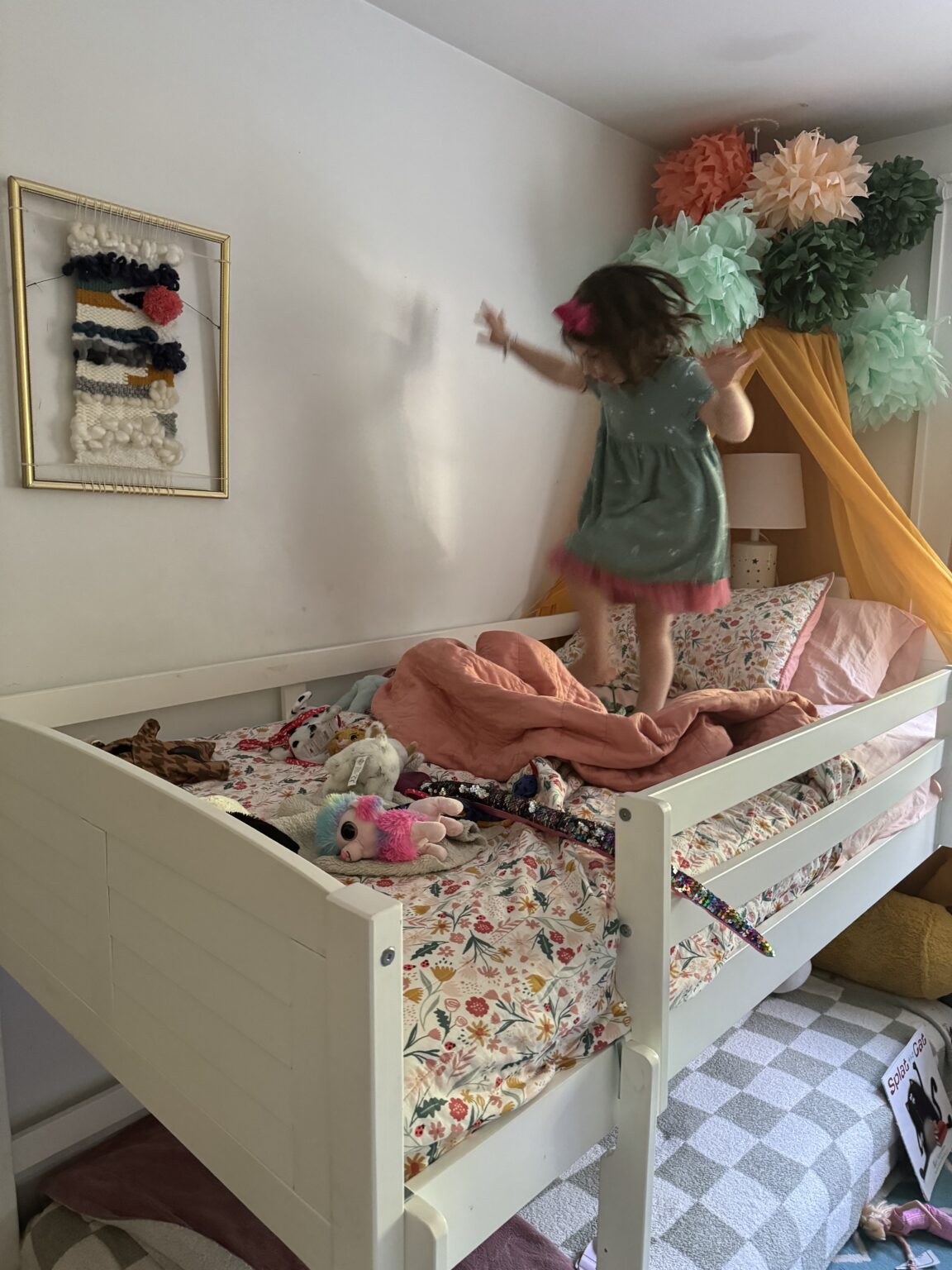 A child in a green dress and pink bow balances on a bed covered with a floral blanket. A canopy with pom-poms hangs above. The room has a textured wall hanging and a patterned rug.