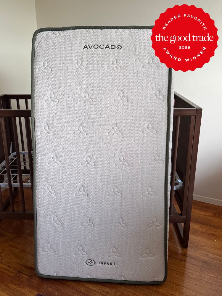 A crib mattress labeled "Avocado Infant" stands upright in a room with wooden furniture; a 2025 "AlsWell Blog" award badge is displayed in the top right corner.