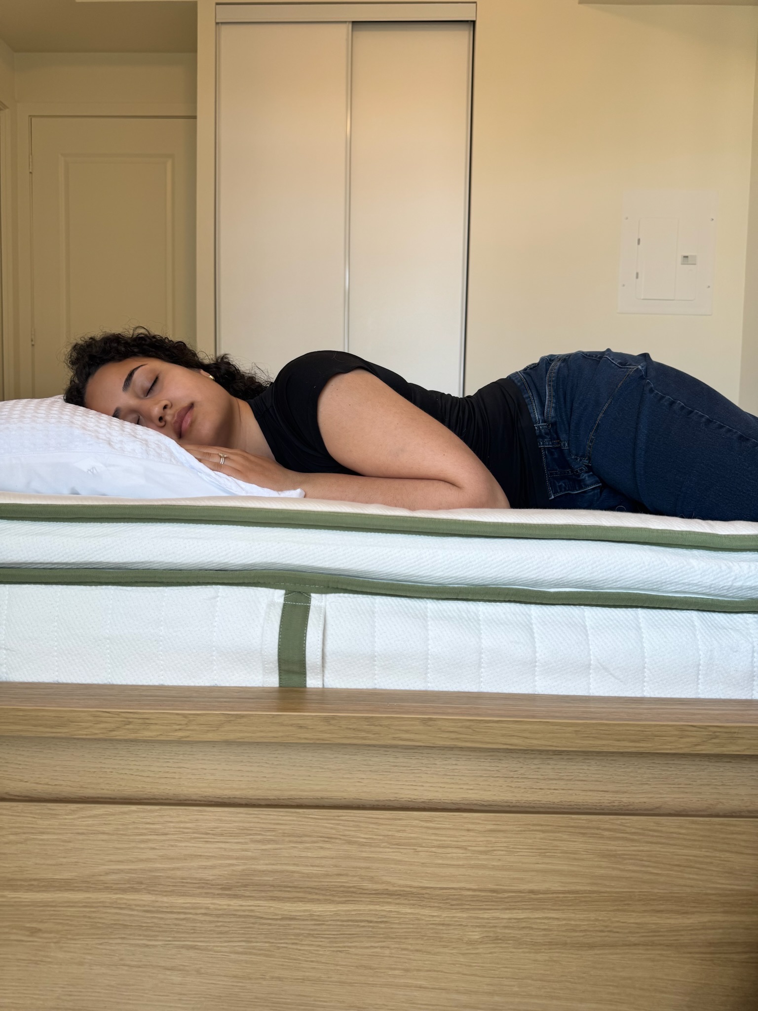 A woman lies on her side on a bed, resting her head on a white pillow with her eyes closed in a bedroom setting.