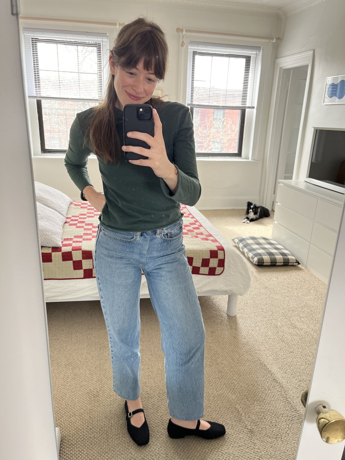 Woman taking a mirror selfie in a bedroom with a red and white quilt on the bed; a black and white cat is lying on a cushion in the background.