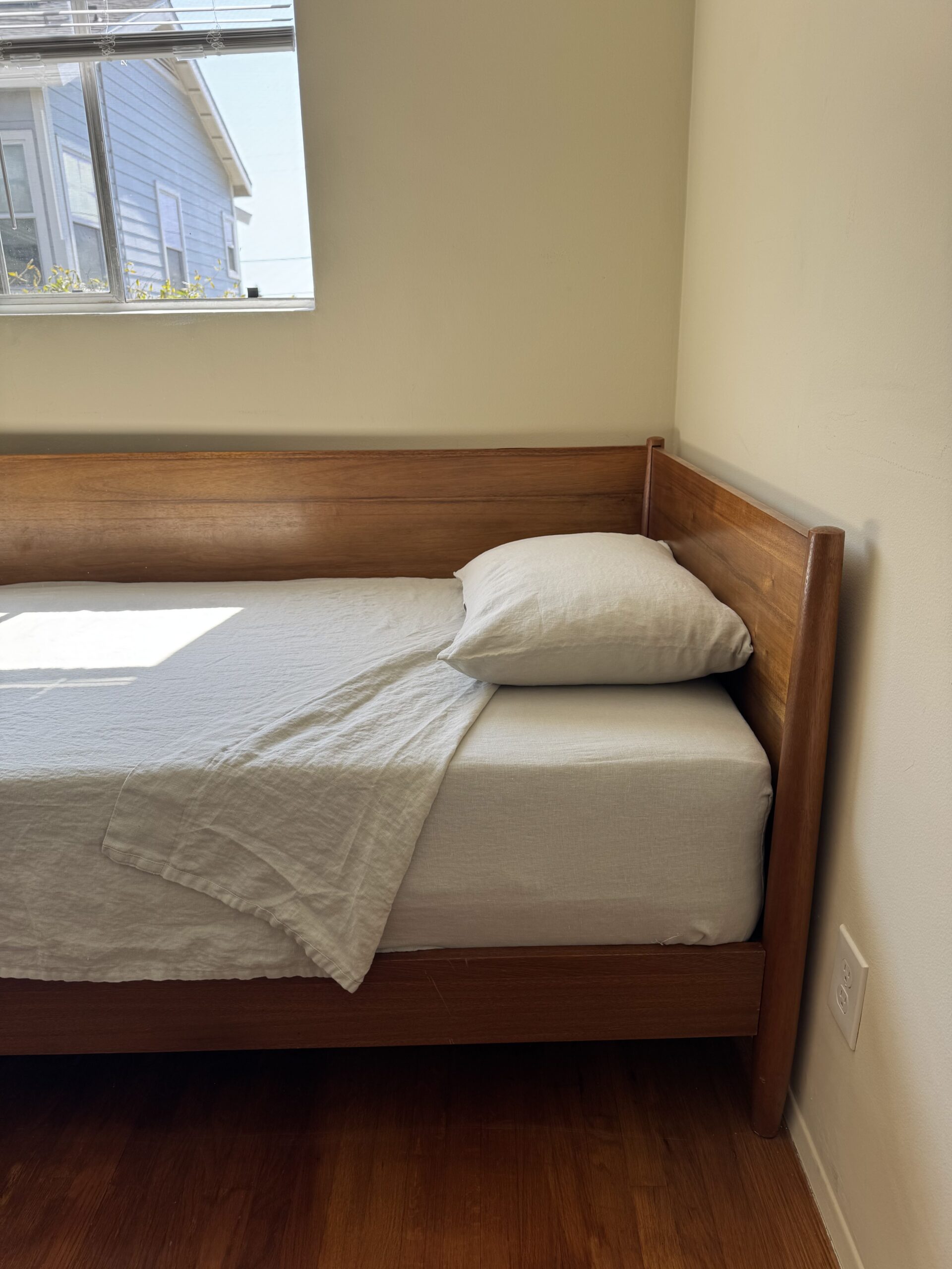 A wooden bed with white bedding and a single pillow is in a corner of a room with light-colored walls and a window. Hardwood floor is visible.