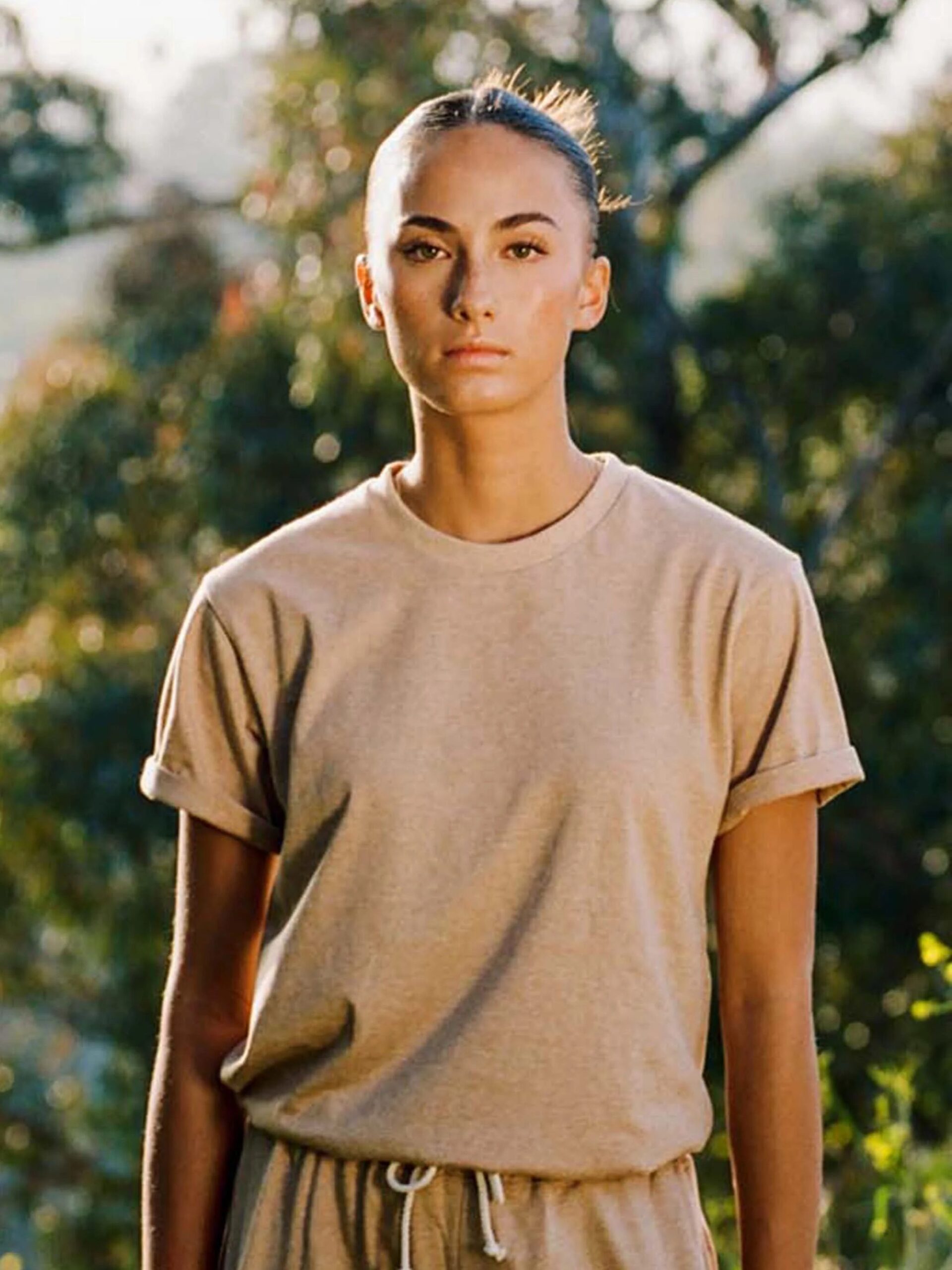 A person in a tan shirt stands outdoors in front of blurred greenery, looking directly ahead.
