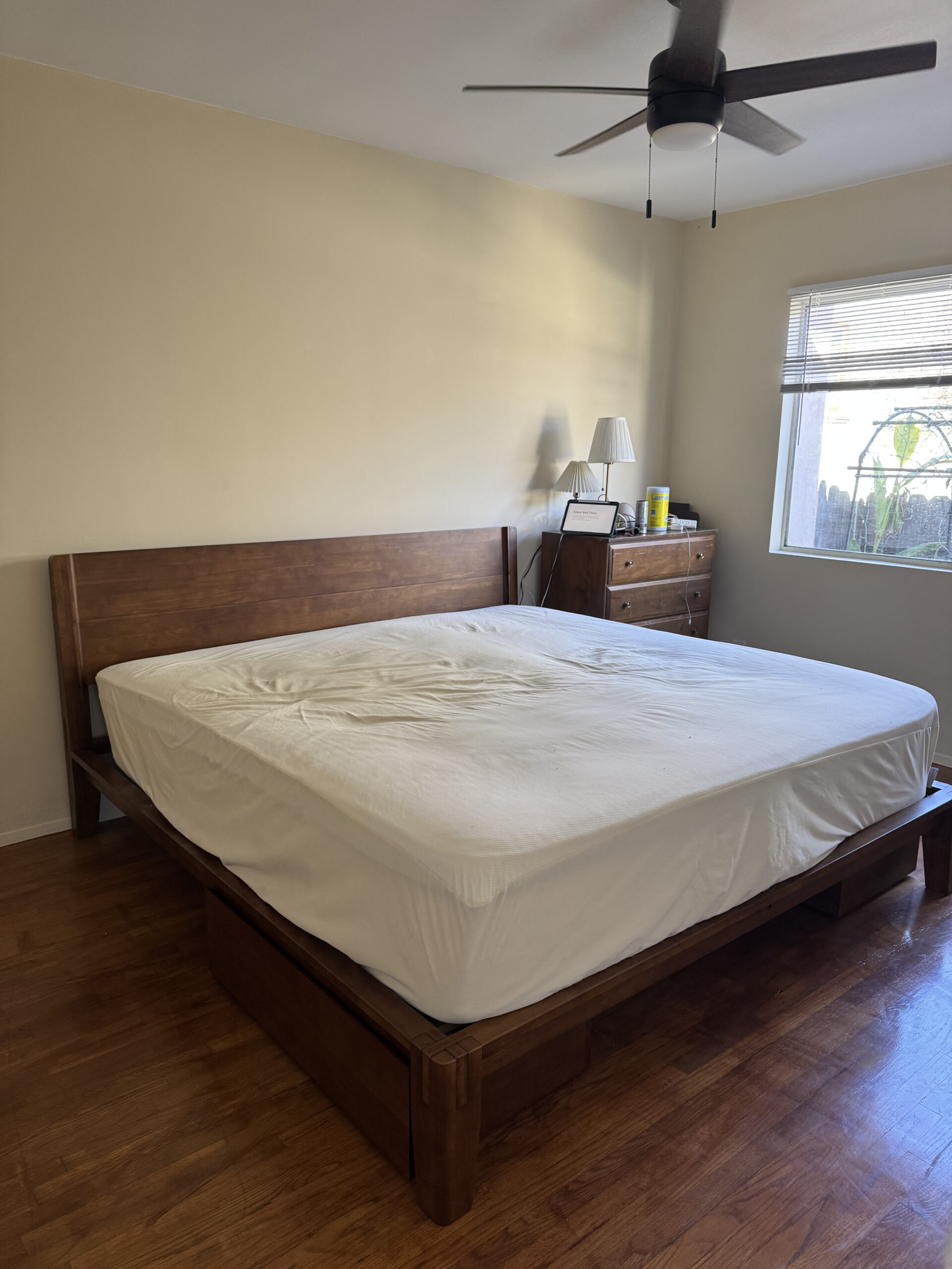 Bedroom with a wooden bed frame and white bedding, a wooden dresser with a lamp and items, wooden floor, ceiling fan, and window with blinds partially open.