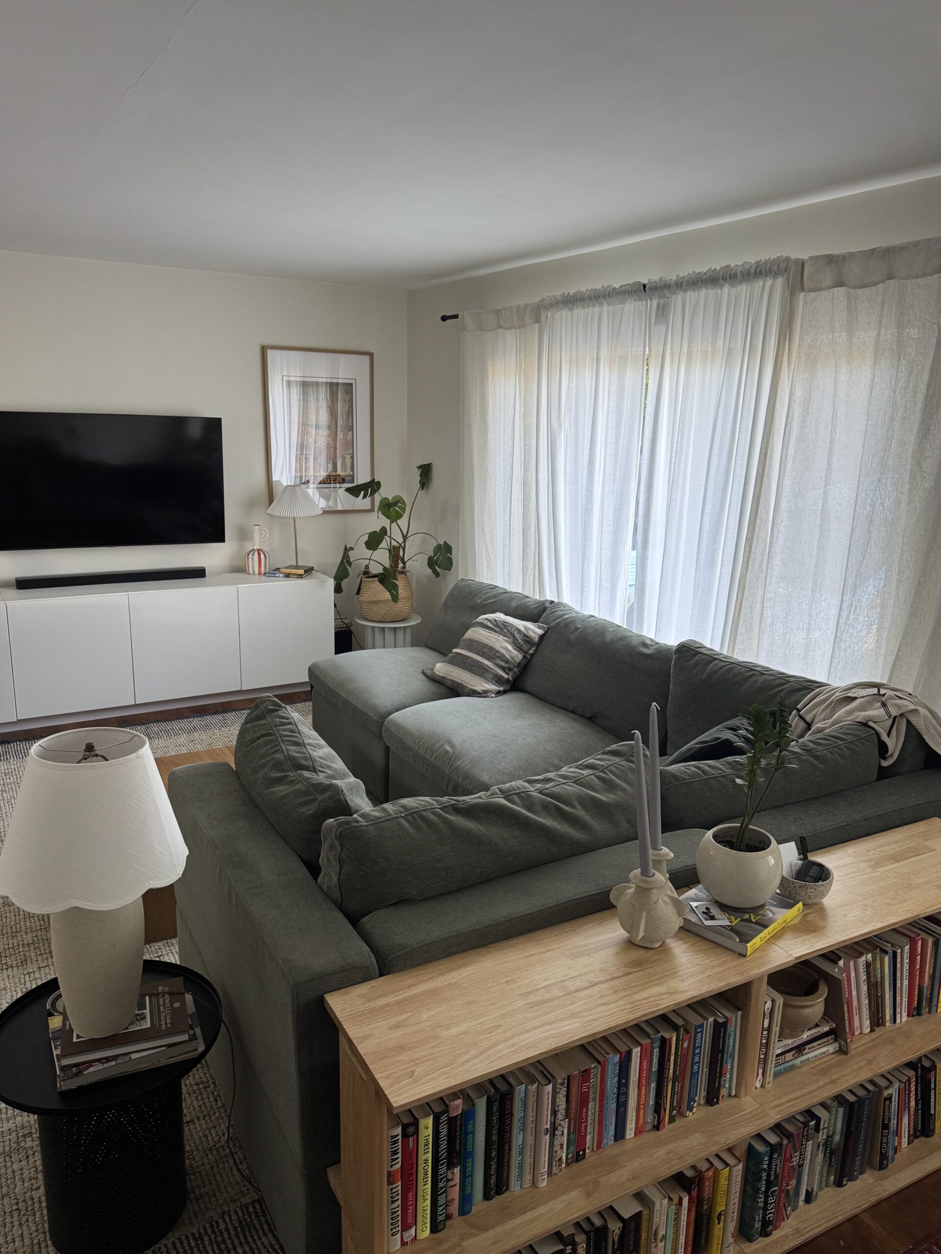 A living room with a green sectional sofa, bookshelf, coffee table, potted plants, large window with sheer curtains, and a wall-mounted TV above a white cabinet.