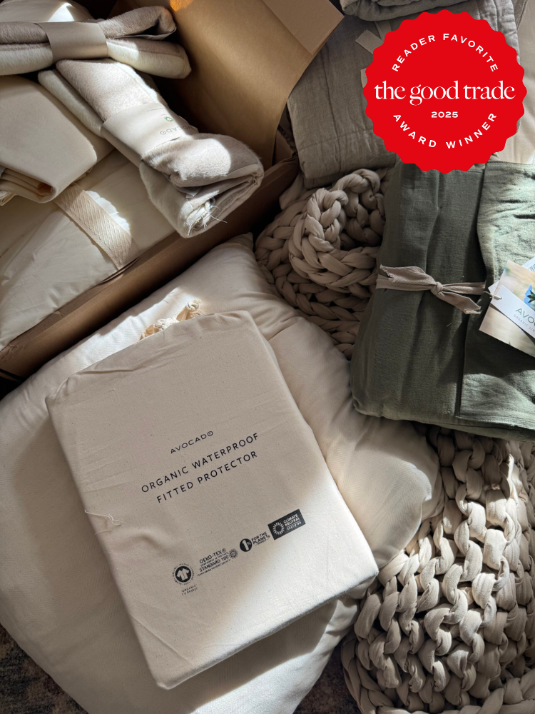 A variety of folded bedding items, including an Avocado organic waterproof fitted protector, displayed on a soft, textured surface with a red "AlsWell Blog Award Winner 2025" badge.