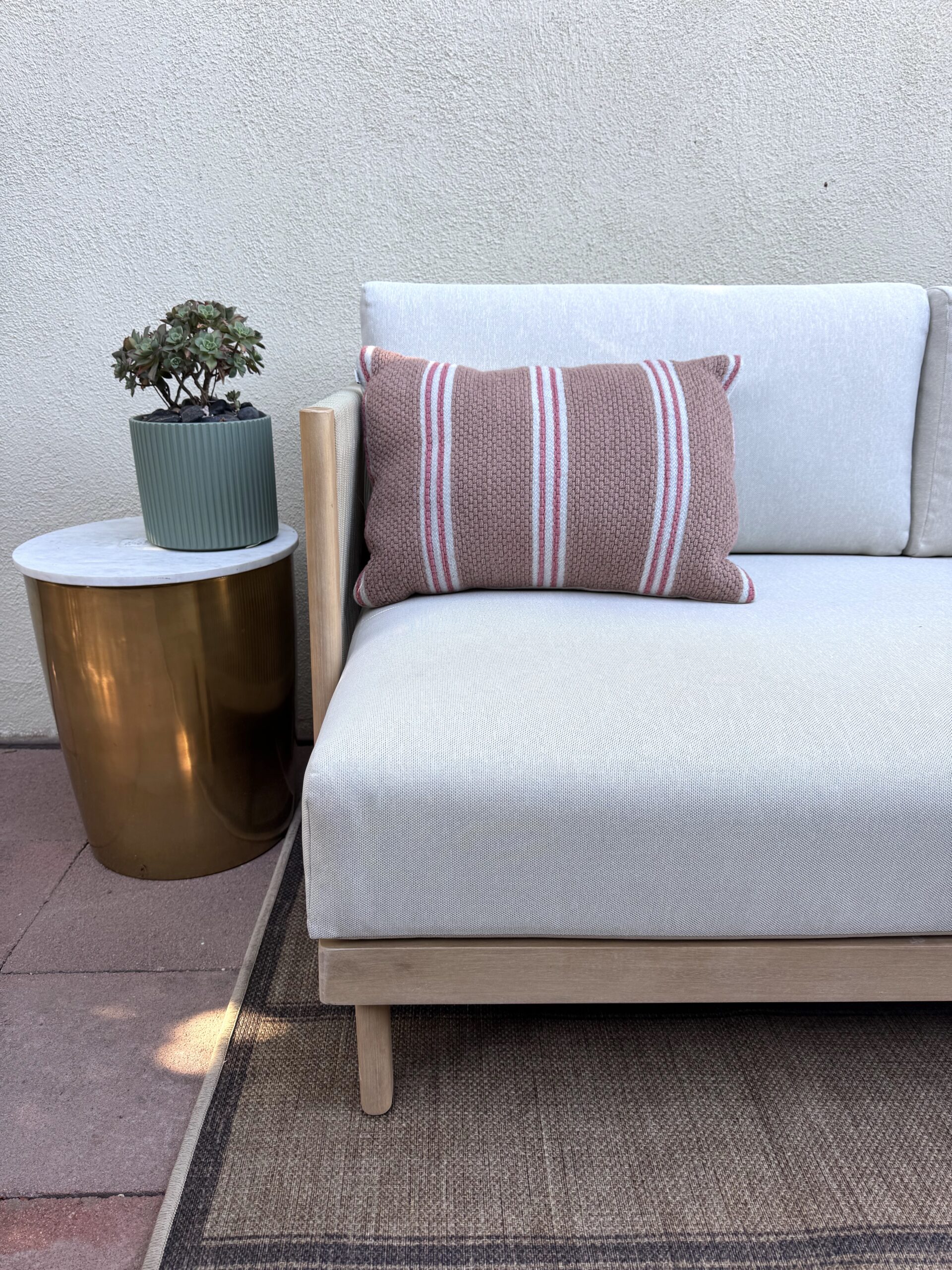 A light-colored outdoor sofa with a striped cushion is next to a gold side table holding a potted succulent, set on a patio with stone tiles and a woven rug.
