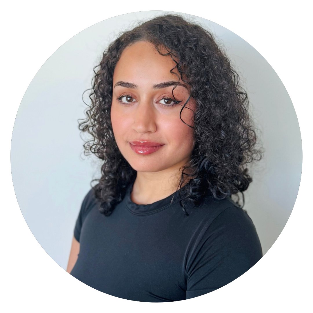 A person with curly dark hair and light skin wearing a black shirt, posing in front of a plain light-colored background, sits comfortably on a latex mattress.